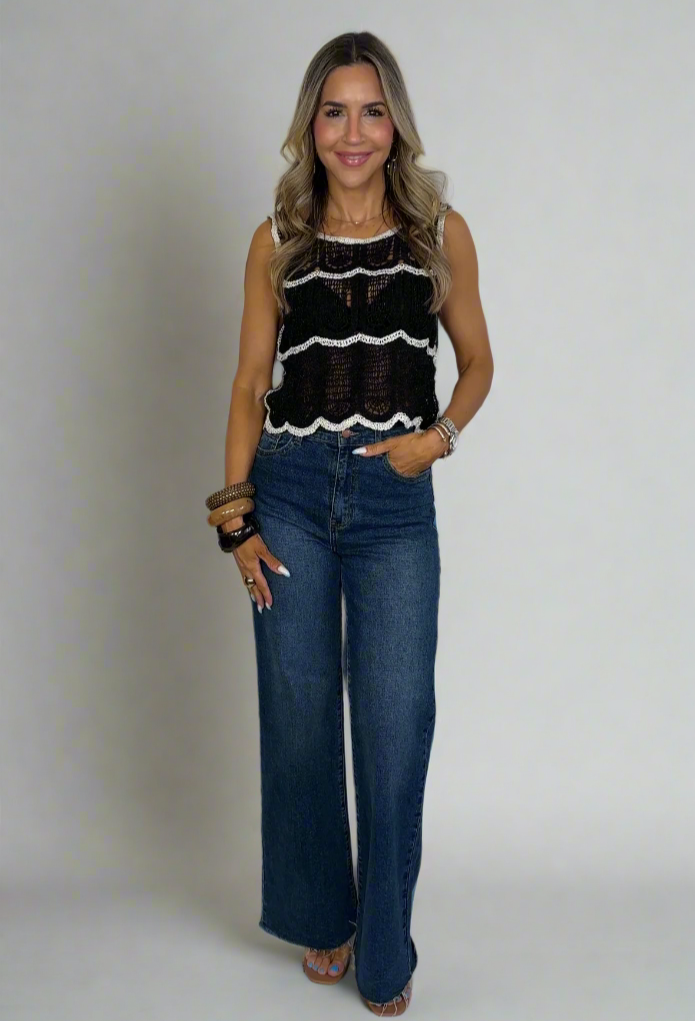 Woman wearing a black sleeveless top with white patterns and blue jeans against a white background